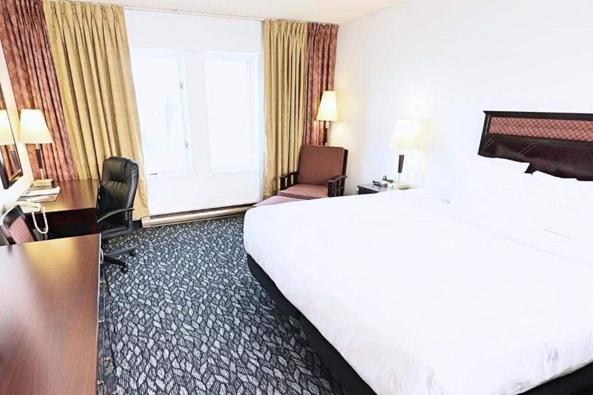 A hotel room with a large bed, a desk, a chair, two lamps, and a view of the outdoors through a window with curtains.