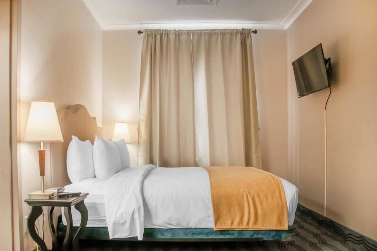 A hotel room with a bed, a nightstand, a lamp, a television mounted on the wall, and a window with curtains.