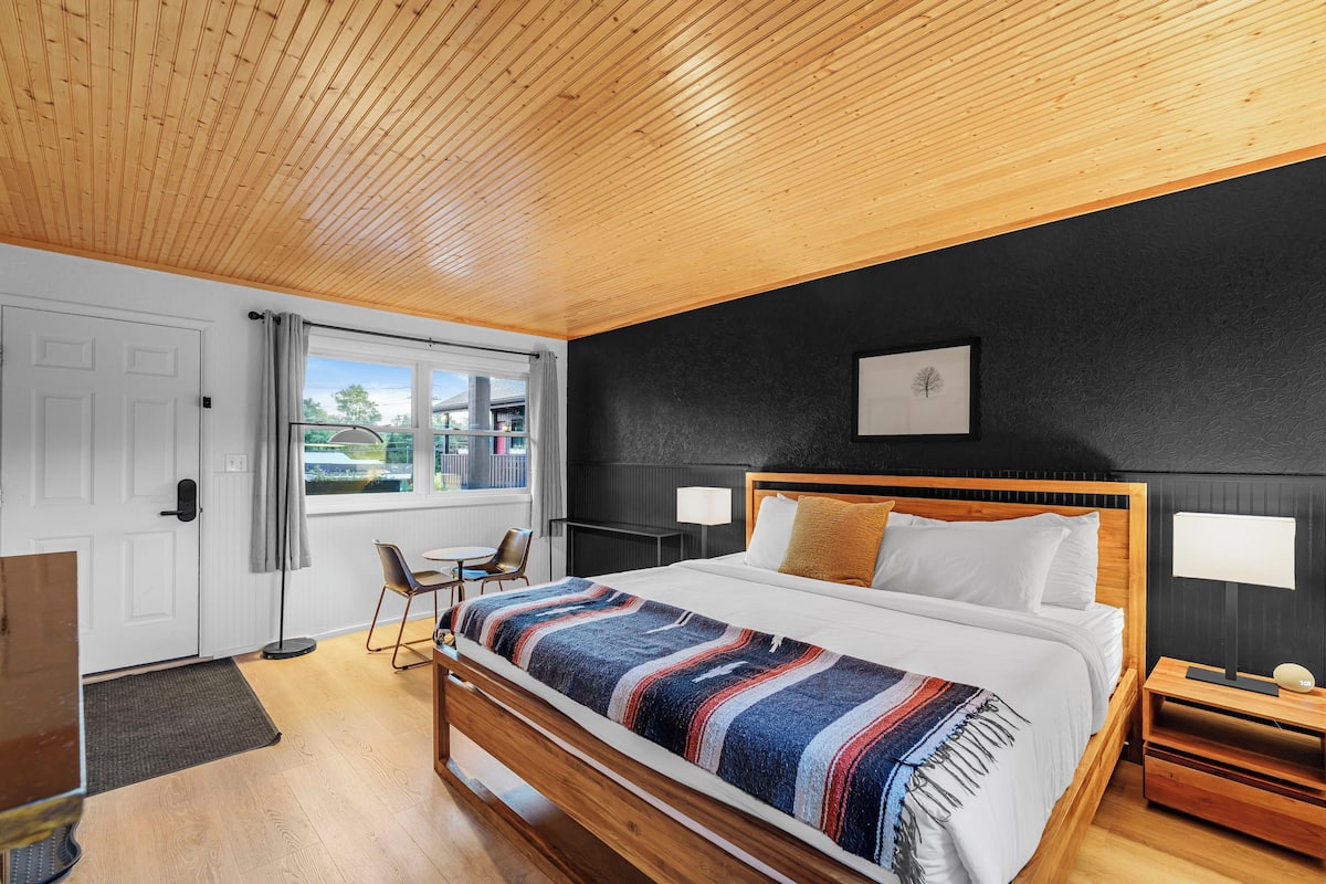 Een moderne slaapkamer met een houten plafond, een groot bed met een kleurrijke deken, een nachtkastje met een lamp en uitzicht op de omgeving door een raam.