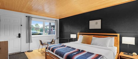 A modern bedroom with a wooden ceiling, a large bed with a colorful blanket, a bedside table with a lamp, and a view of the outdoors through a window.
