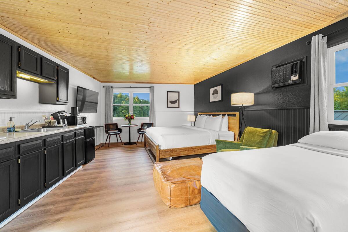 Een moderne hotelkamer met een houten plafond, een kitchenette, twee bedden, een zithoek en een groot raam met uitzicht op groen.