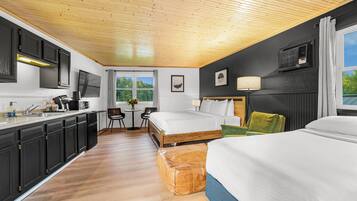 A modern hotel room with a wooden ceiling, a kitchenette, two beds, a dining area, and a large window with a view of greenery.
