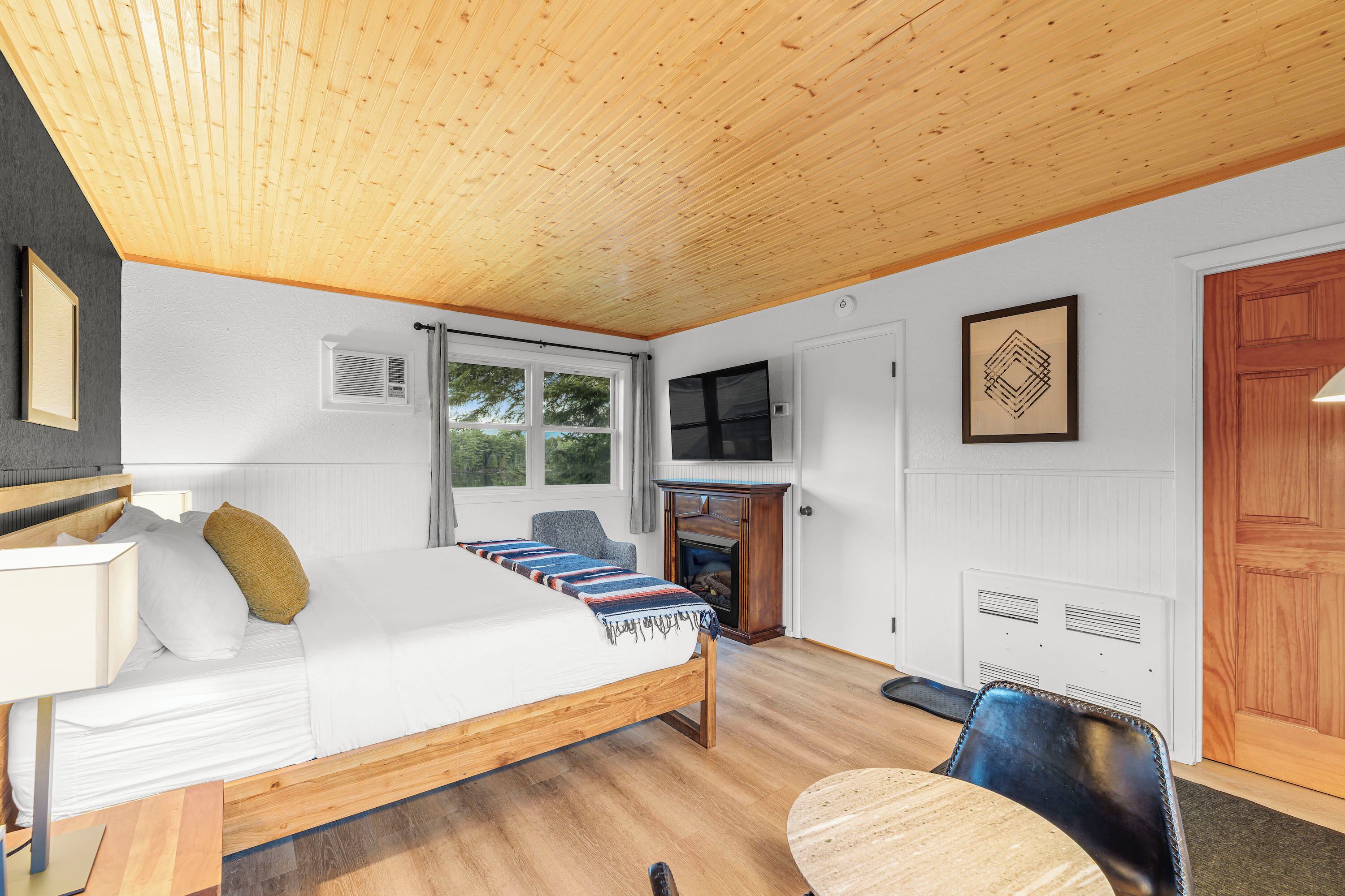 Een slaapkamer met een houten plafond, een bed met een kleurrijke deken, een televisie, een open haard en een deur.