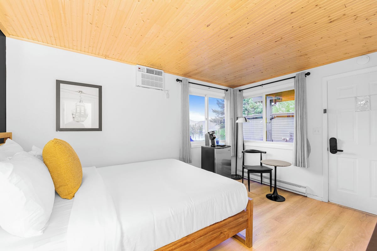 Een slaapkamer met een houten plafond, een bed met wit en geel beddengoed, een klein tafeltje en een stoel.