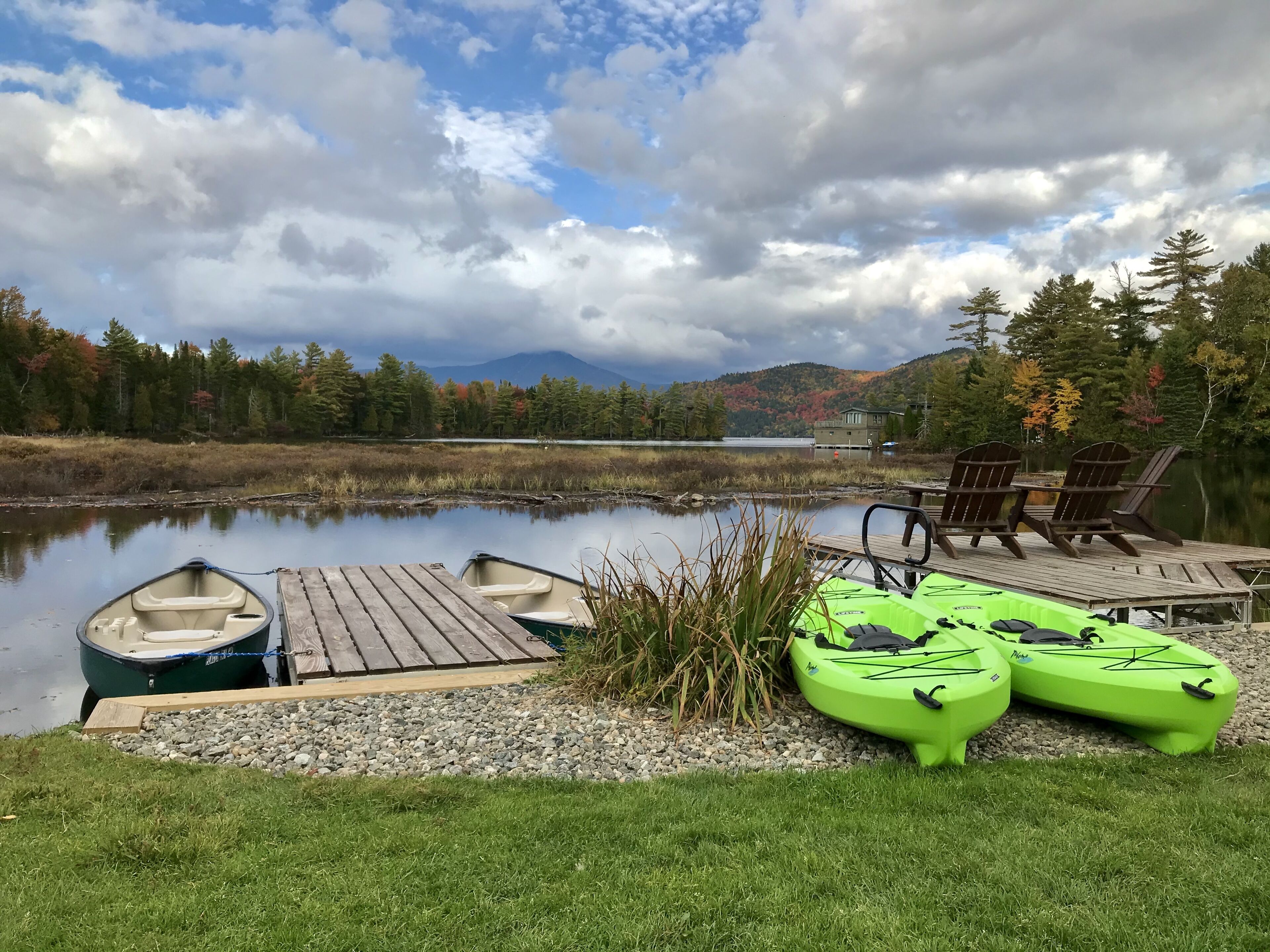 Un muelle a orillas del lago con un bote y kayaks, rodeado de árboles y una montaña al fondo.