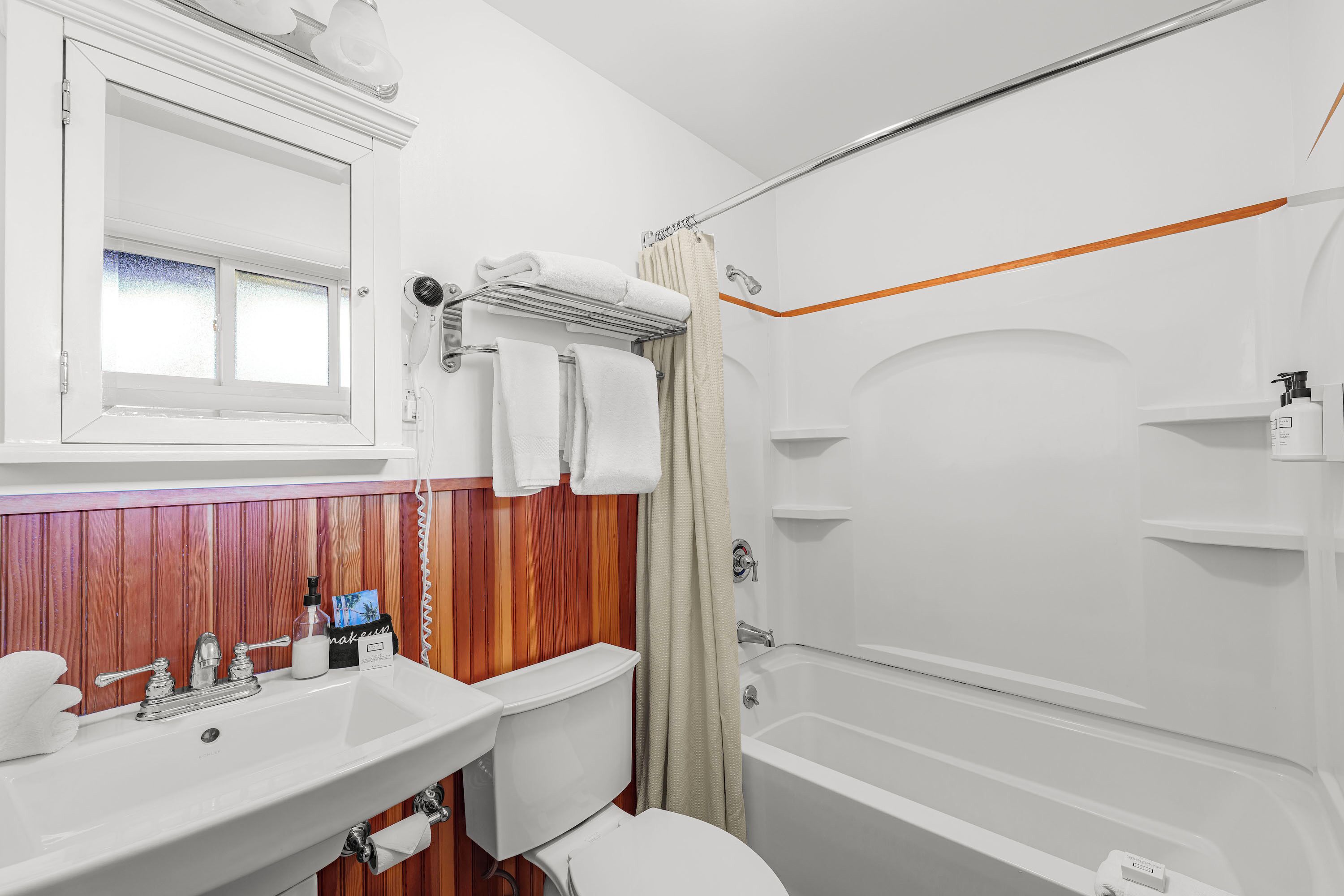 Un baño con un lavabo blanco, inodoro y bañera. Hay una ventana sobre el lavabo, y un perchero con toallas.