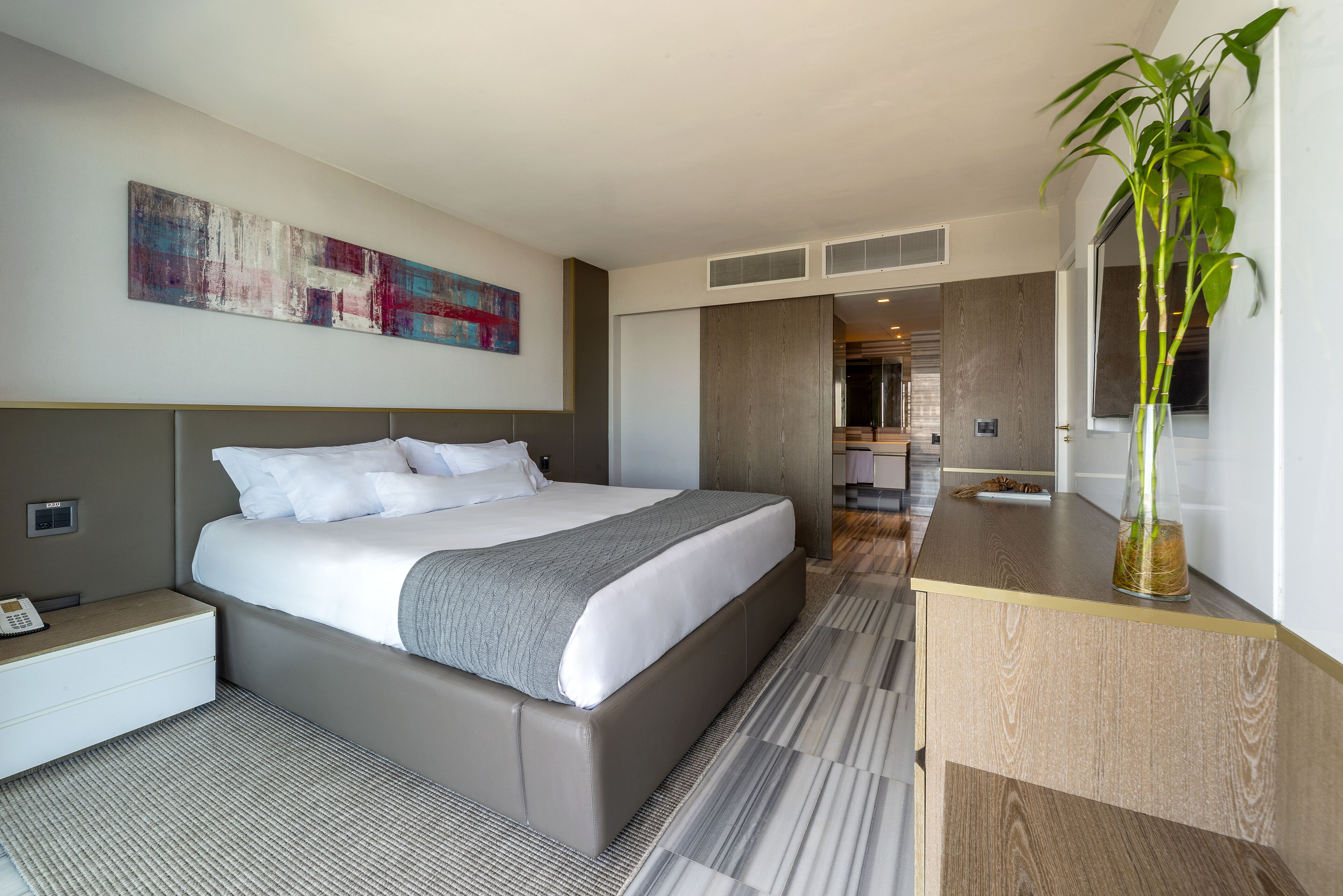 A modern hotel room with a large bed, a bedside table, a nightstand, a desk, and a potted plant.