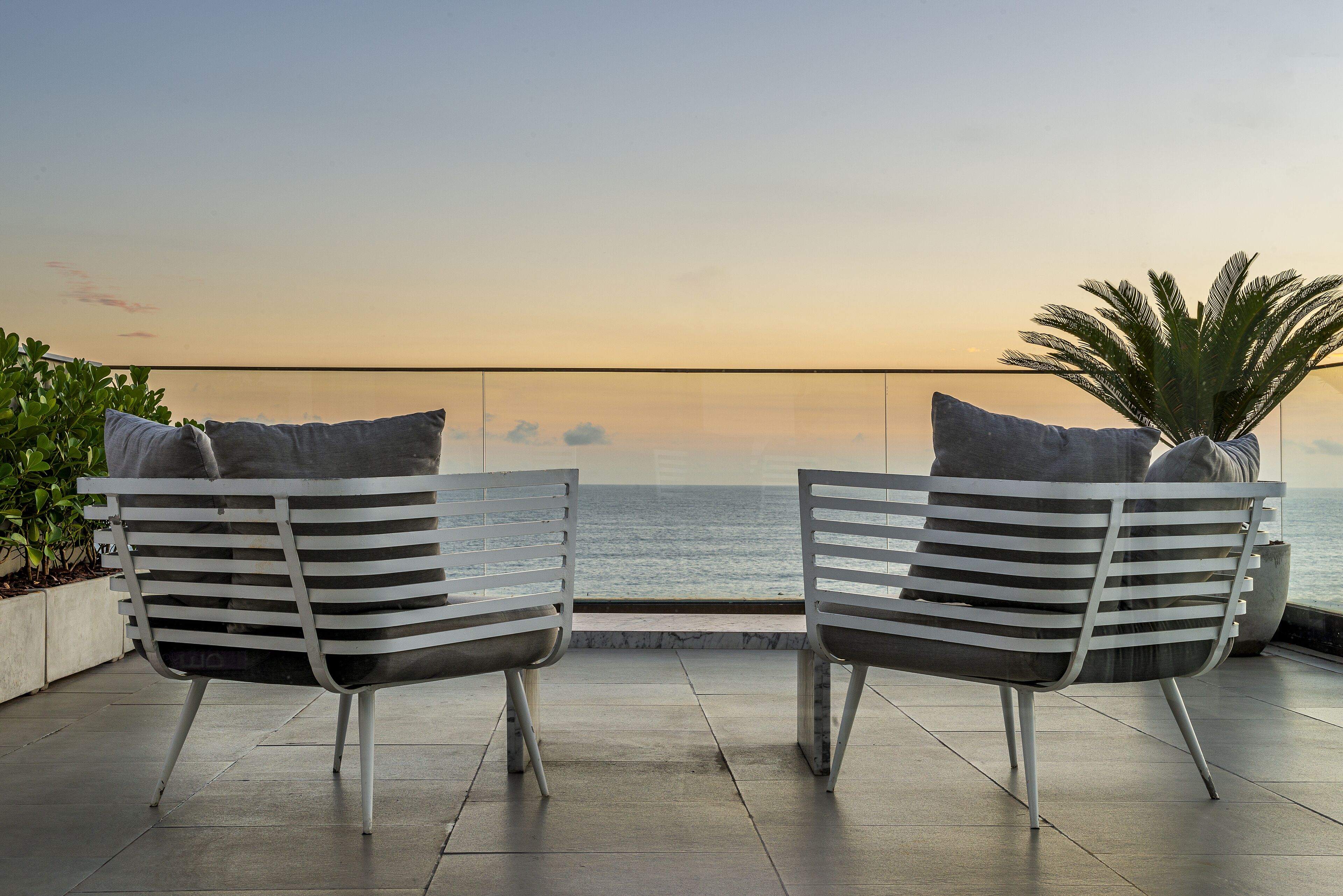 Two white chairs with grey cushions facing a balcony with a view of the sea.