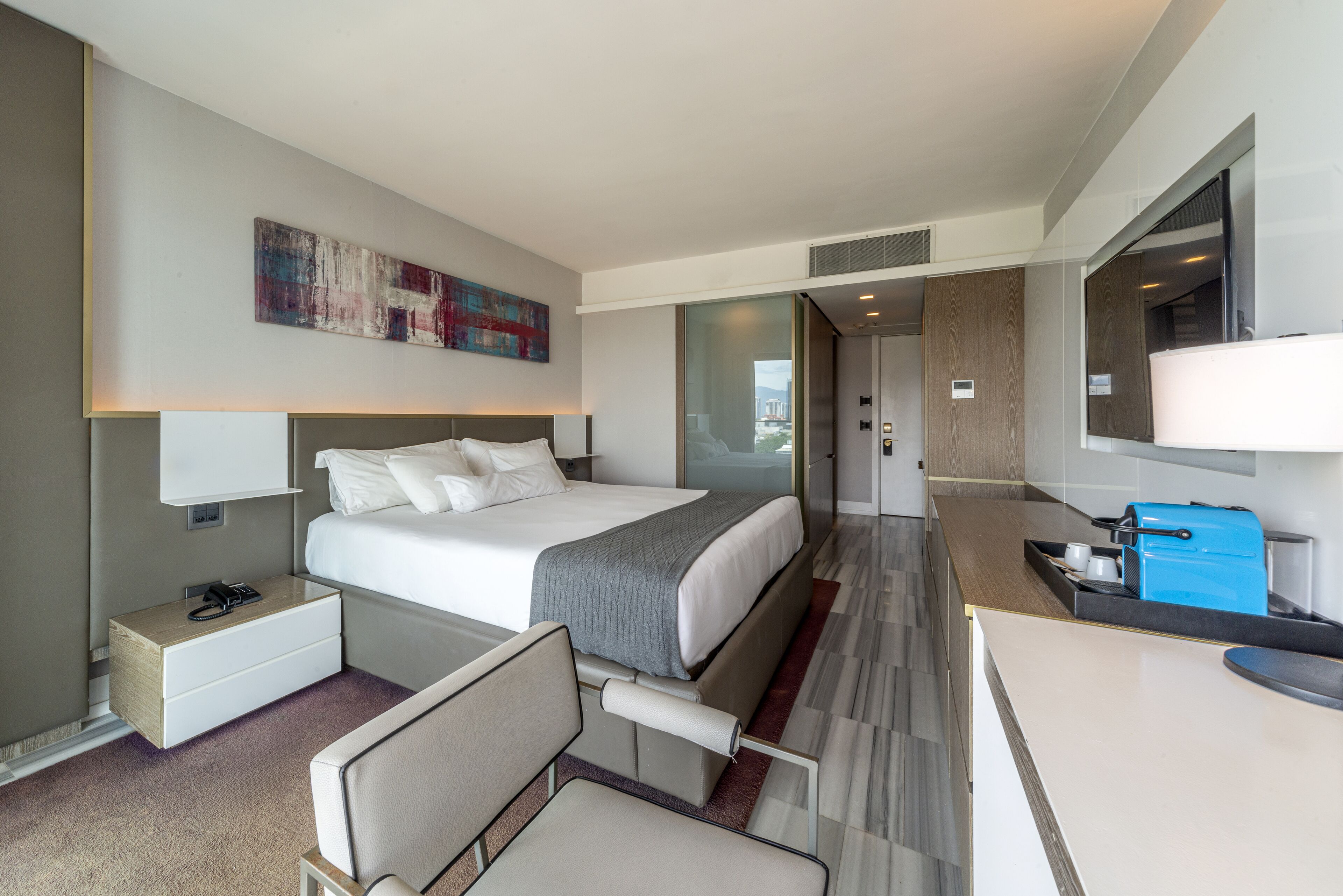 A modern hotel room with a bed, bedside table, chair, and a small kitchenette area.