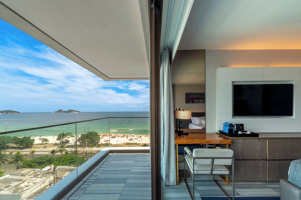 Quarto moderno com uma grande janela com vista para a praia, uma TV de tela plana na parede e uma escrivaninha de madeira com uma cadeira.