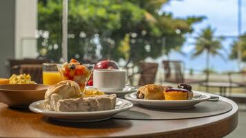 A breakfast table with various dishes including pastries, fruit, and a cup of coffee, set against a view of palm trees and the ocean.