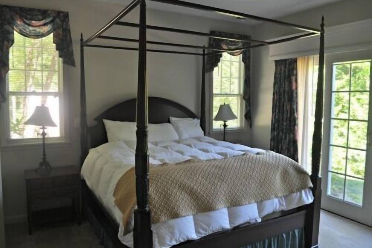 Una cama con dosel, ropa de cama blanca, estructura de madera oscura y una manta de color beige. Hay dos mesitas de noche con lámparas, y la habitación tiene ventanas grandes con cortinas.