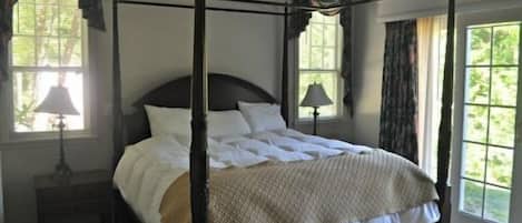 A four-poster bed with white linens, a dark wooden frame, and a beige throw blanket. There are two bedside tables with lamps, and the room has large windows with curtains.