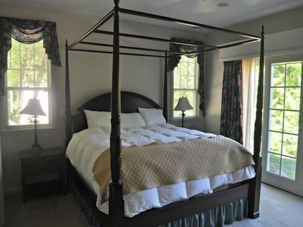 A four-poster bed with white linens, a dark wooden frame, and a beige throw blanket. There are two bedside tables with lamps, and the room has large windows with curtains.
