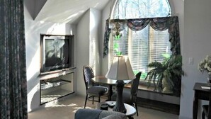 Una sala de estar con televisión, una mesa de comedor, sillas, una lámpara y un ventanal grande con cortinas.
