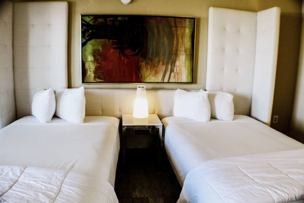 Habitación de hotel con dos camas, una mesita de noche con lámpara y un cuadro abstracto grande en la pared.