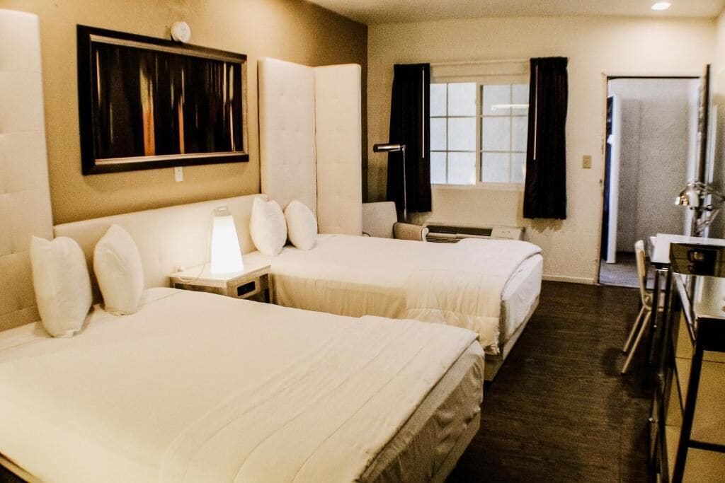 A hotel room with two beds, a bedside table, a lamp, a window with curtains, and a door.