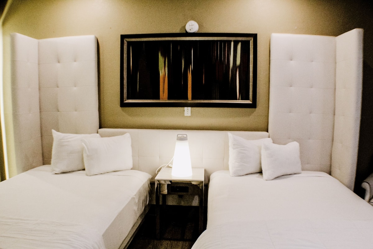 Habitación de hotel con dos camas, una mesita de noche, un reloj en la pared y una ventana con persianas.