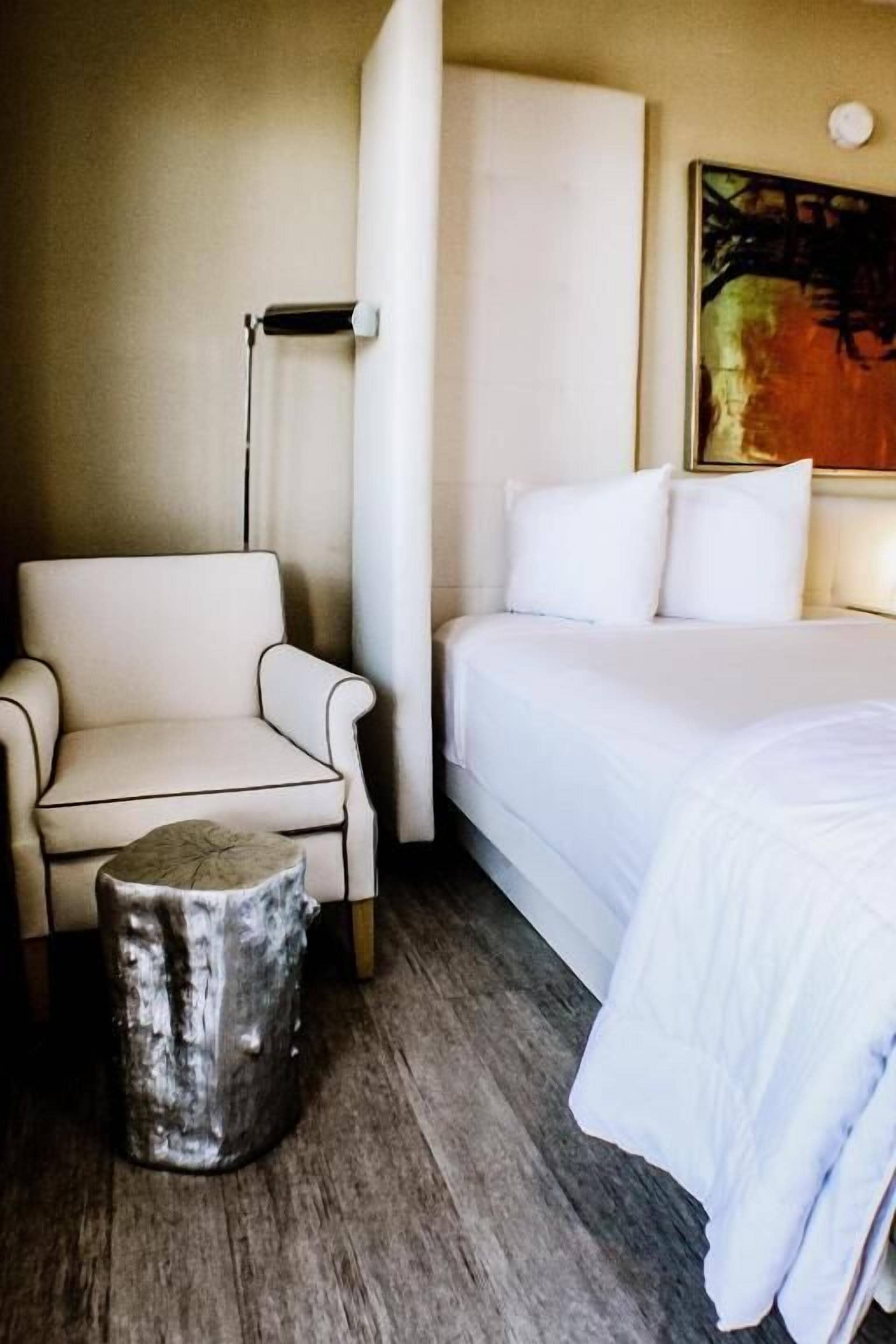 A hotel room with a bed, a chair, a small wooden stool, and a painting on the wall.