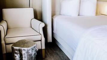 A hotel room with a bed, a chair, a small wooden stool, and a painting on the wall.