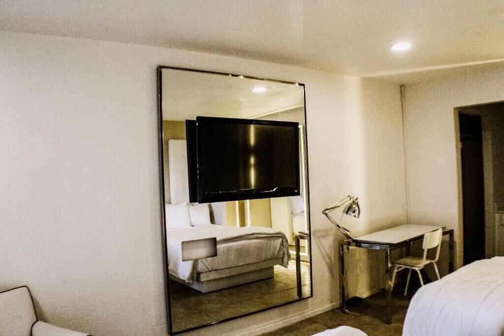 A hotel room with a large mirror reflecting a bed, a desk with a chair, and a television.
