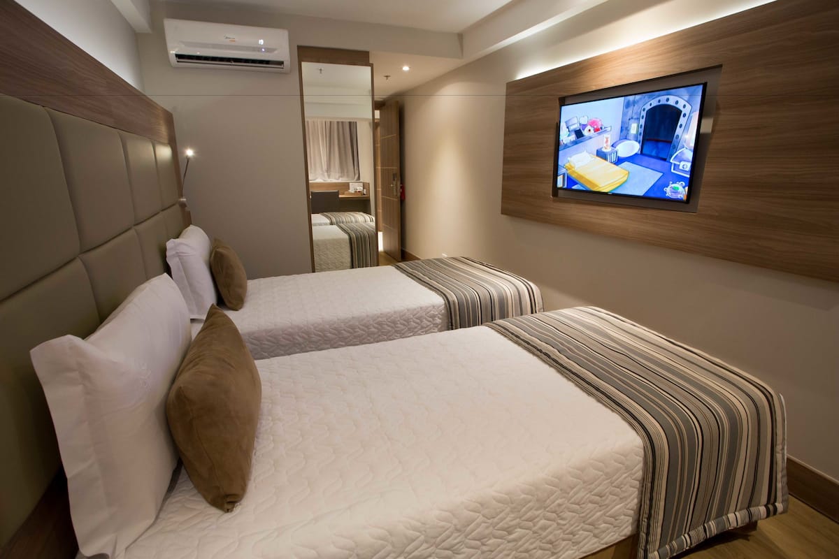 Quarto de hotel com duas camas, uma televisão fixada na parede e um ar condicionado.