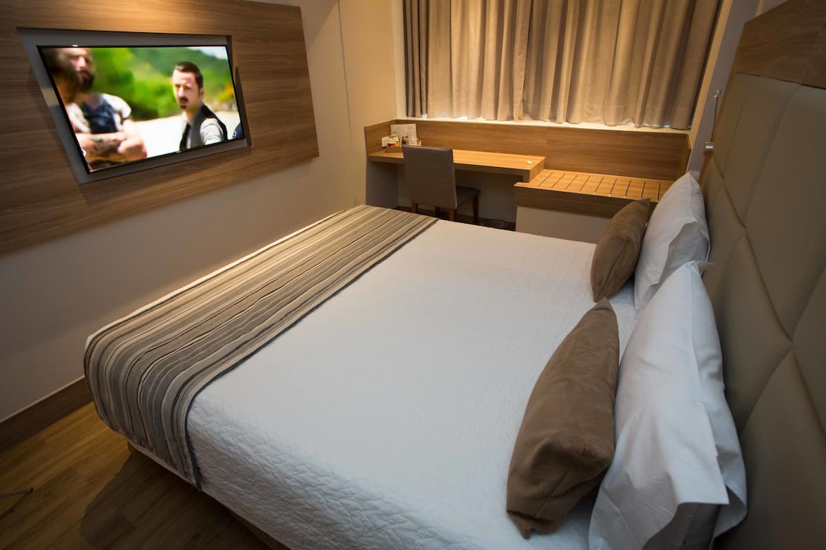 Quarto de hotel com cama, escrivaninha e televisão fixada na parede.