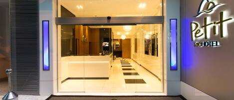 The entrance of Petit Rio Hotel with a clear glass facade, a reception desk, and a modern interior visible through the glass.