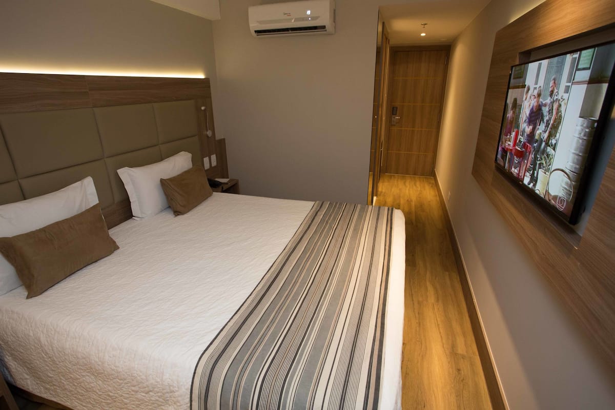 Quarto de hotel com uma cama grande, televisão de tela plana fixada na parede e um aparelho de ar condicionado acima da cama.
