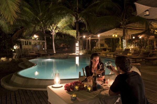 Una pareja cenando junto a una piscina por la noche, con una mesa adornada con velas y una botella de vino.