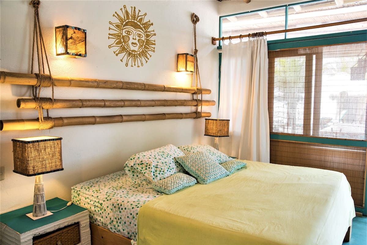A bed with a green and white polka dot bedspread, two pillows, and a bedside lamp. There is a wall decoration of a sun and a window with a curtain.