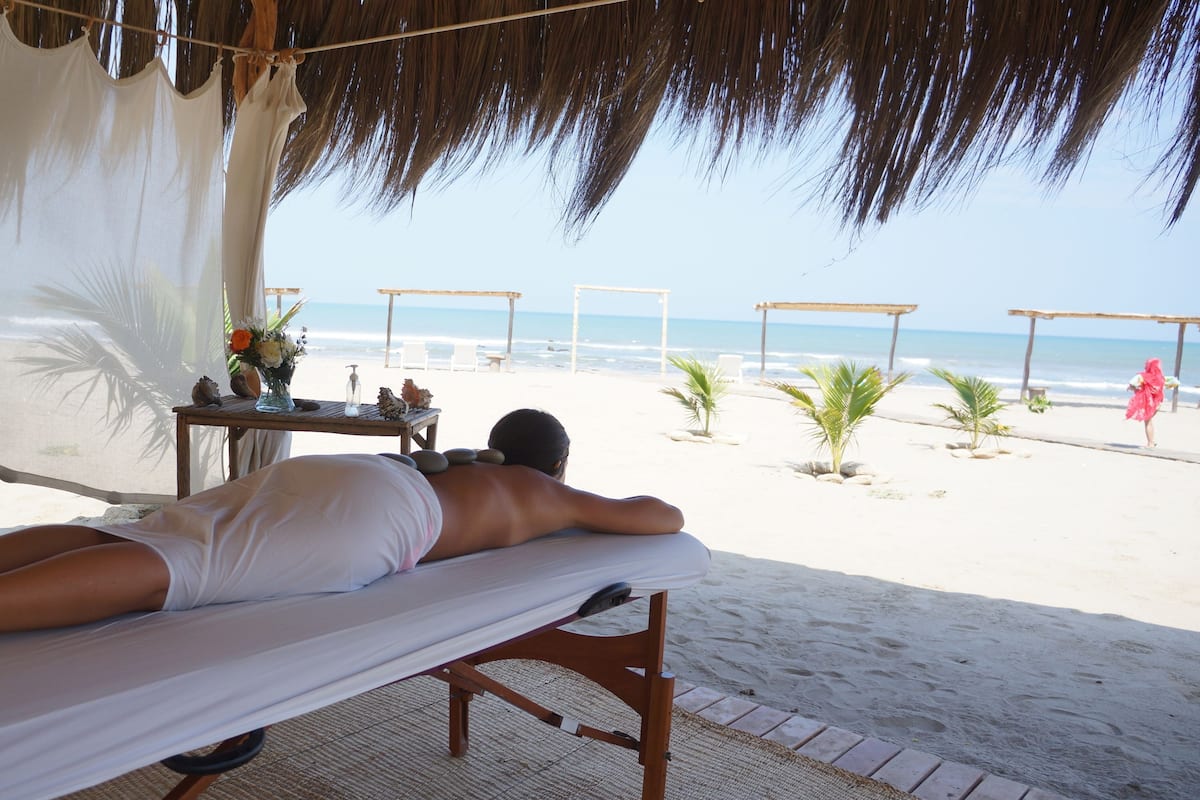 A beachside massage area with a person lying on a massage table, a table with a vase of flowers, and a view of the beach and ocean.