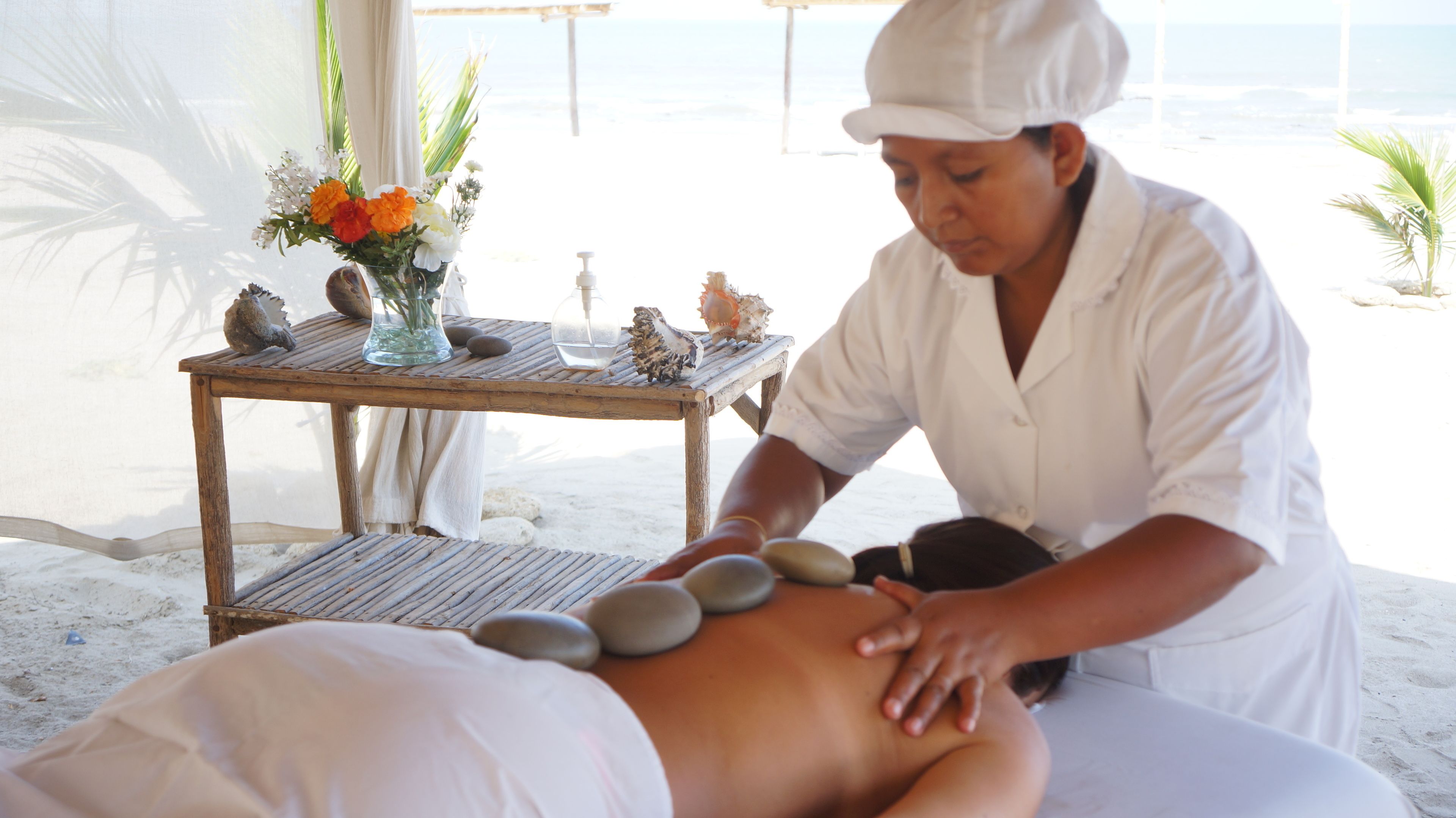 Une séance de massage sur une table en bois, agrémentée de pierres et de fleurs, avec vue sur une plage.