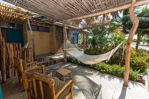 Un área de estar al aire libre con muebles de madera, una hamaca y vistas a plantas tropicales y un edificio al fondo.