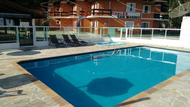 En swimmingpool med solstolar, en byggnad med en skylt där det står ”CHALÉS SAN MARINO”, och en klar himmel.