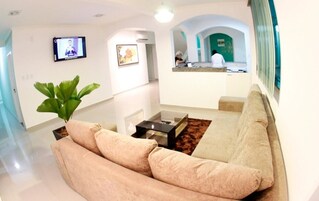 Uma sala de estar moderna com um sofá bege, uma mesa de centro e uma planta grande. Uma televisão está montada na parede, e uma área de cozinha é visível ao fundo.