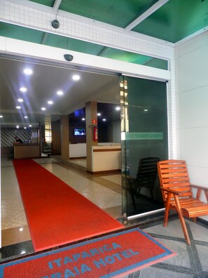 A hotel reception area with a red carpet leading to the entrance, a reception desk, and a glass door with a sign indicating an automatic fire alarm system. - Itaparica Praia Hotel (Vila Velha)