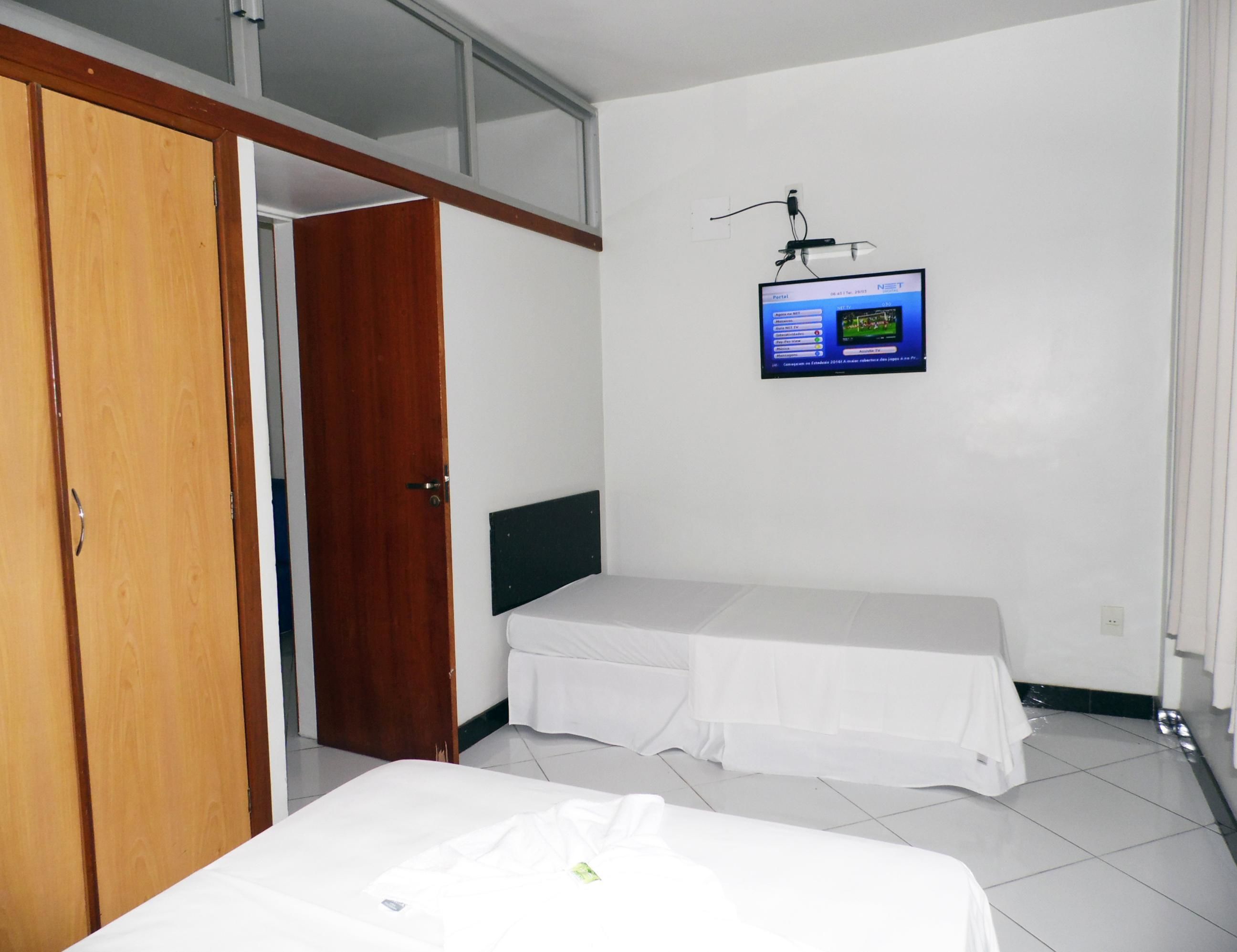 Quarto com guarda-roupa de madeira, cama com colchão branco e cabeceira preta, pequena mesa de cabeceira e TV fixada na parede exibindo um jogo.