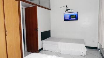 Quarto com guarda-roupa de madeira, cama com colchão branco e cabeceira preta, pequena mesa de cabeceira e TV fixada na parede exibindo um jogo.