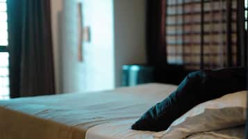 A bed with a white sheet and a dark pillow, a metal trash bin, and a window with blinds.