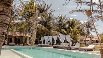 A pool area with lounge chairs and a cabana, surrounded by palm trees and a building with a tiled roof.