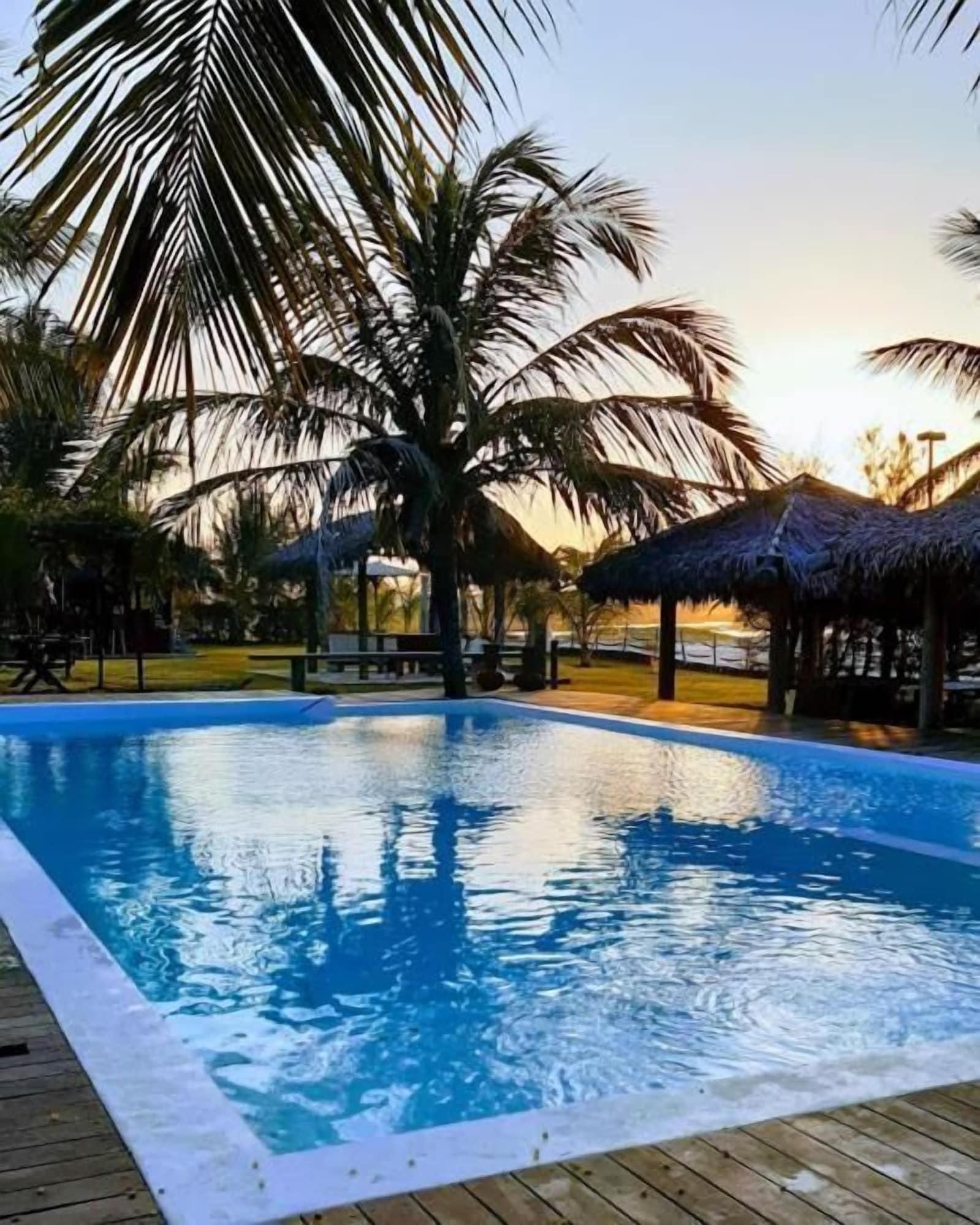 Piscina com deck de madeira, cercada por palmeiras e estruturas com telhado de palha.
