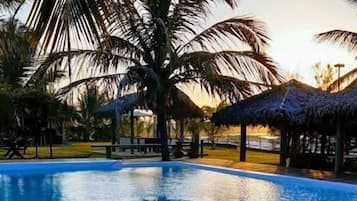 Piscina com deck de madeira, cercada por palmeiras e estruturas com telhado de palha.
