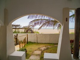 Vista através de uma porta arqueada para uma praia, com uma estrutura de telhado de palha e palmeiras ao fundo.