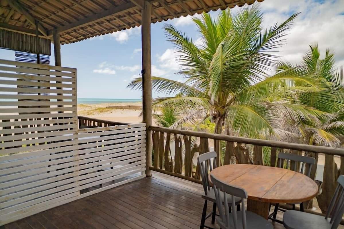 Una terraza de madera con una mesa redonda y sillas, con vistas a una playa con palmeras y un cielo despejado.