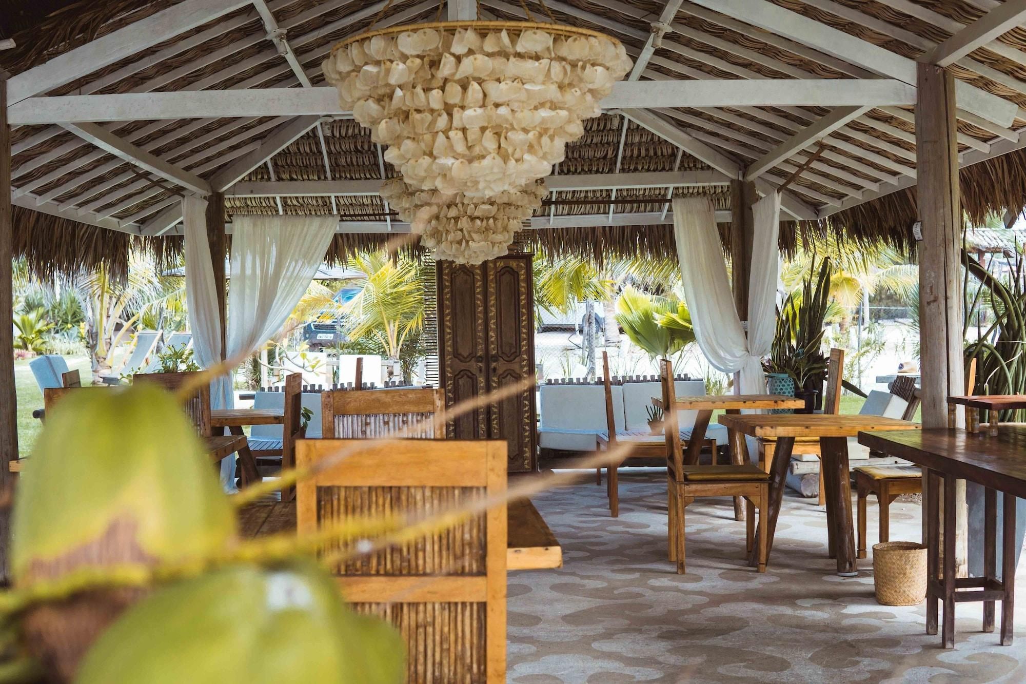 An outdoor dining area with wooden tables and chairs, a thatched roof, and a hanging light fixture.