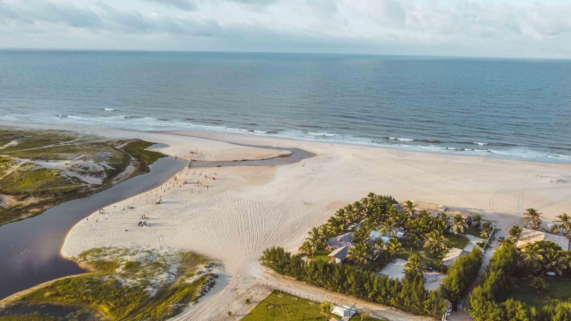 Vista aérea de uma praia com costa curvilínea, uma pequena área de água e um conjunto de edifícios com palmeiras.