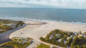 An aerial view of a beach with a curved shoreline, a small water body, and a cluster of buildings with palm trees. - Zebra Boutique Hotel (Beberibe)