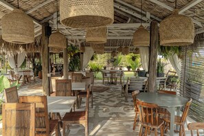 Un espace de restauration en plein air avec des tables et des chaises en bois, des lampes suspendues en fibres tressées et un sol à motifs.