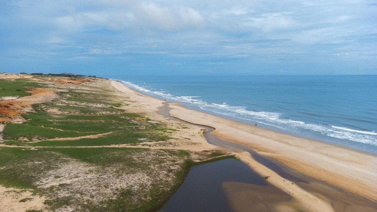 Vista aérea de uma praia com céu claro, faixa de areia e uma pequena área de água.